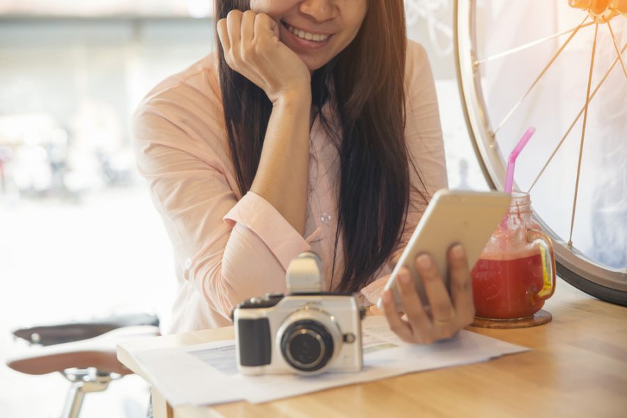 Beautiful young woman using smart phone. She is smiling and happy.On the wooden table is a camera (mirrorless) and fruit juice (watermelon).